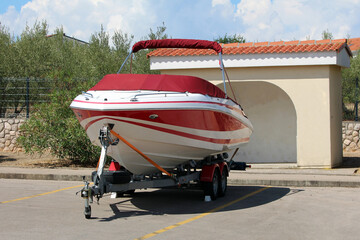 A sleek red and white fiberglass speedboat is tightly strapped to a double-axle trailer, parked in...