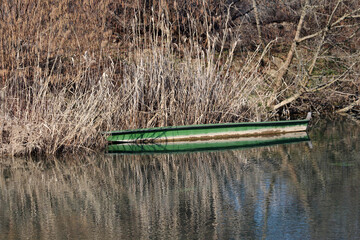 A weathered green wooden boat rests quietly along a calm riverbank, partially hidden by dry reeds...