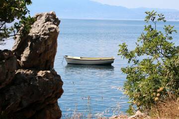 A white rowboat drifts peacefully on a calm lake, framed by rugged rocks and leafy branches, with...