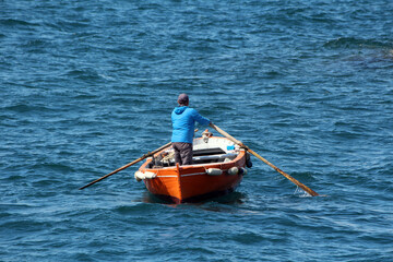 A lone person in a blue jacket rows a vivid orange wooden boat with long oars across rippling blue water, creating gentle waves and evoking a sense of peaceful isolation and adventure