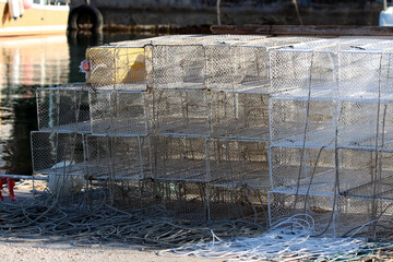 A large pile of rectangular wire fishing traps is stacked near the water’s edge, with tangled...