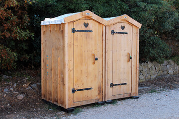 Two side-by-side wooden outdoor toilets feature heart-shaped ventilation cutouts on their doors,...
