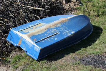 A faded blue rowboat with peeling paint and rust stains lies overturned on a grassy bank, showing...