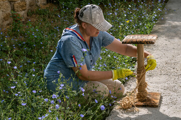 Woman in denim dress and baseball cap repairs cat scratching post outdoors, symbolizing pet care...