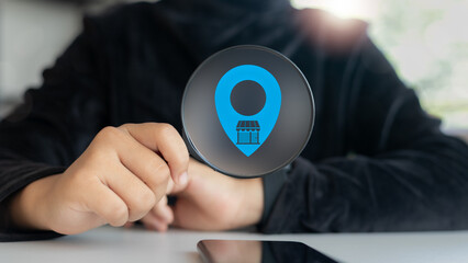 Close-up hand holding a magnifying glass with a blue map pin and a store icon. Concept of local...