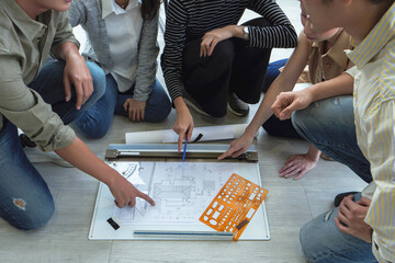 Group of young Southeast Asian architects and designers collaborating on blueprint floor plan, pointing at details during creative meeting on floor workspace.