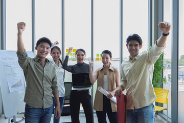 Group of young Southeast Asian business professionals raising fists in celebration inside modern office with large windows. Team smiling confidently after project success.