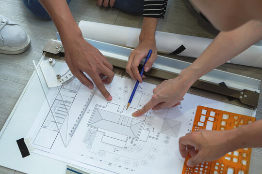 Close-up view of Southeast Asian architect team collaborating on detailed building blueprint using pencil and scale ruler in modern workspace.