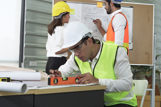 Asian engineer in safety helmet and reflective vest using spirit level to measure blueprint on desk, with colleagues discussing project plan in background at modern construction office. - Powered by Adobe