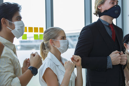 Diverse business team wearing protective face masks standing indoors showing determined fists, expressing unity and motivation during challenging times