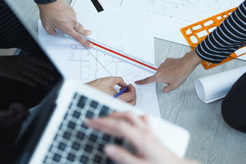 Close-up of architecture team reviewing blueprint on floor, pointing at details of construction plan with ruler and template, captured from top-down perspective in natural light.