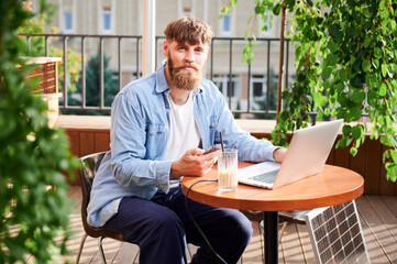 Man works on laptop and smartphone at outdoor table, with glass of iced coffee. Solar panel nearby,...