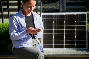 Happy woman using smartphone connected to photovoltaic solar panel. Integration of sustainable...