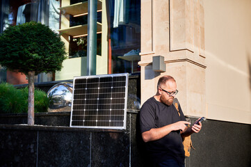 Bearded man using smartphone next to photovoltaic solar panel. Integration of sustainable renewable...