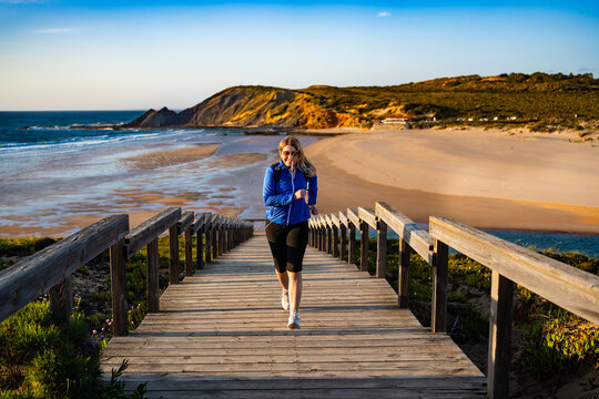 Beautiful mid adult woman running up wooden stairs at viewpoint on Atlantic ocean and 
Amoreira beach in Portugal on spring evening. Front view	