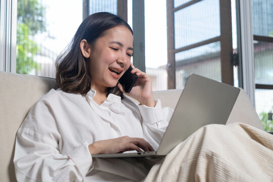 A young woman smiles while talking on her phone and using a laptop, seated comfortably in a bright, modern room with large windows. - Powered by Adobe