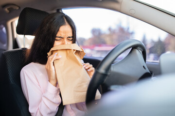 Indian woman breathing into paper bag inside car, showing signs of anxiety or panic. Concept of...