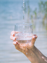 a man holds a glass of clear water in his hand against the backdrop of a forest lake