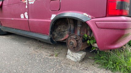 A faded, magenta-colored car is partially visible, with a detached, rusted wheel supported by...
