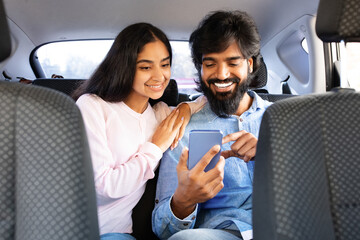 Young smiling indian couple sitting in car backseat, using smartphone together. Shows intimacy, relaxation and digital lifestyle during modern car travel, closeup