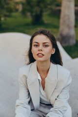 Woman in light grey business suit sitting outdoors with confident expression, natural background,...