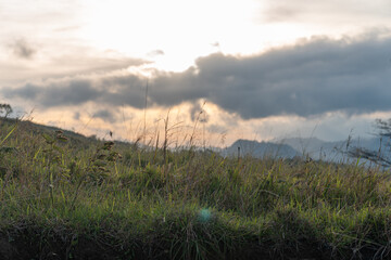 Peaceful sunset scene with grassy hillside, layered mountains, and dramatic clouds. Soft light and natural colors create a calm and atmospheric landscape view.