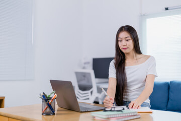 Focused Asian businesswoman sitting at a desk, taking notes and working on a laptop in a home office, embodying remote work and a flexible freelance lifestyle