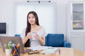 Beautiful Asian businesswoman enjoying coffee while working on a laptop at home, embodying the work-from-home lifestyle with a blend of comfort and modern technology