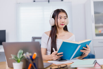 Asian female student wearing headphones, reading a book, and taking notes while engaging in online...