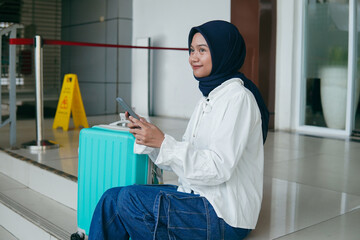 Portrait of Asian hijab woman resting on train station stairs, using mobile phone, waiting for...