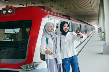 Two young Muslim women in hijab taking a selfie together in front of a train, smiling happily before starting their journey