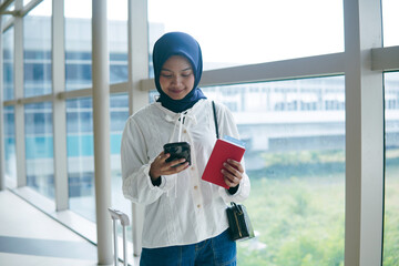 Asian woman in hijab walking through airport corridor, holding passport and smartphone, ready for...