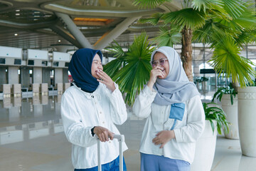 Two Asian women in hijab chatting cheerfully at the airport, enjoying their time together before the flight