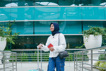 Asian woman in hijab walking through airport corridor, holding her passport and heading confidently...