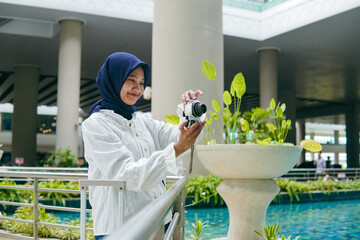 Asian woman in hijab taking photos of the airport atmosphere with a pocket camera, capturing...