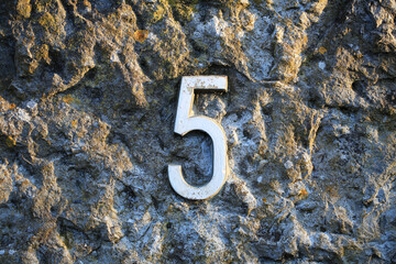 A weathered metal number 5 mounted on a rough, textured stone wall. The surface patina and natural...