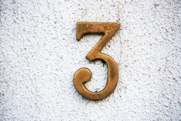 Brass number 3 mounted on a textured white stucco wall. The aged metal shows patina and character, ideal for concepts like signage, address numbers, vintage design, or typography