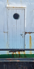 A weathered white boat door with a round porthole shows signs of rust and age, set against a backdrop of peeling paint and a black railing