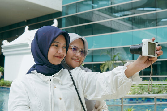 Two Asian women in hijab taking a cheerful selfie with a pocket camera at the airport, capturing memories before their flight