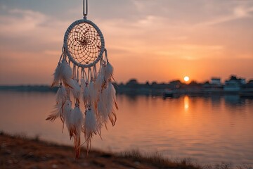 White dreamcatcher with feathers hangs against a serene sunset over calm water and distant buildings