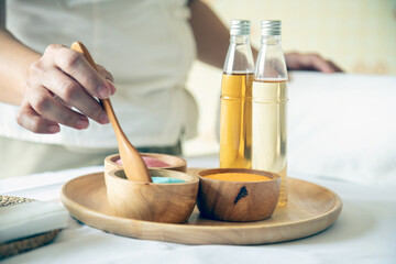 Close-up of hand holding wooden spoon above colorful spa salts in wooden bowls with two glass bottles of essential oil placed on round tray