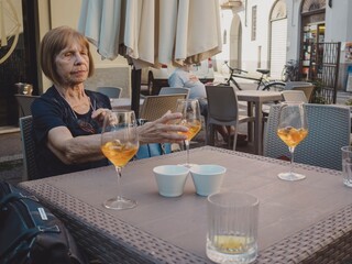 Mature white haired woman sipping citrus cocktail at cafe table, blurred bicycles and patrons...