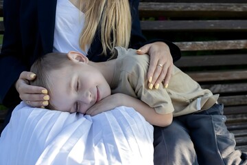 A mother gently holds her child on her lap while sitting on a park bench. The child, looking...