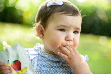 Adorable toddler girl sucking her fingers while holding a fruit puree pouch in a garden. Baby enjoying fruit snack outdoors, showcasing real-life healthy eating habits