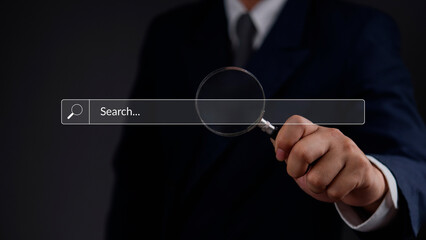 Businessman holding a magnifying glass over a digital search bar, data discovery and information...