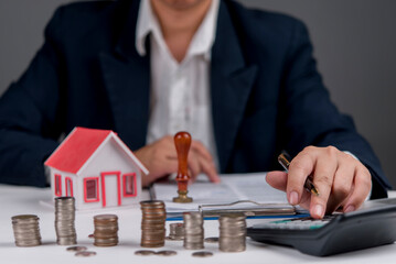 Businessman calculating real estate investment, with house model, coins, and calculator, symbolizing property finance, mortgage planning, home loan, asset growth, financial advisor.