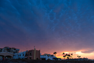 Dramatic sunset sky with mammatus clouds over coastal houses and palm trees, with a bird flying and warm orange and purple tones painting the horizon in a summer evening scene