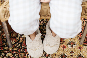 Women's legs in trousers, soft slippers on their feet against the background of a home carpet.