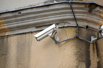 Outdoor security camera mounted on a weathered wall with visible cables, capturing surveillance in an urban environment. The camera shows signs of aging and use.
