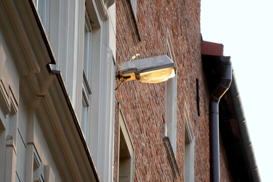 Old street light mounted on a building wall with a weathered yellowed glass cover. The lamp is attached to a brick wall with visible electrical wires and a few bird spikes on top to prevent perching.  - Powered by Adobe